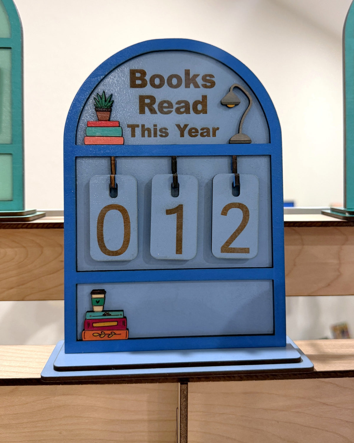 Souvenir Design Books Read This Year wooden counter in blue, number tiles showing 012, with a coffee cup and stacked books on the bottom shelf