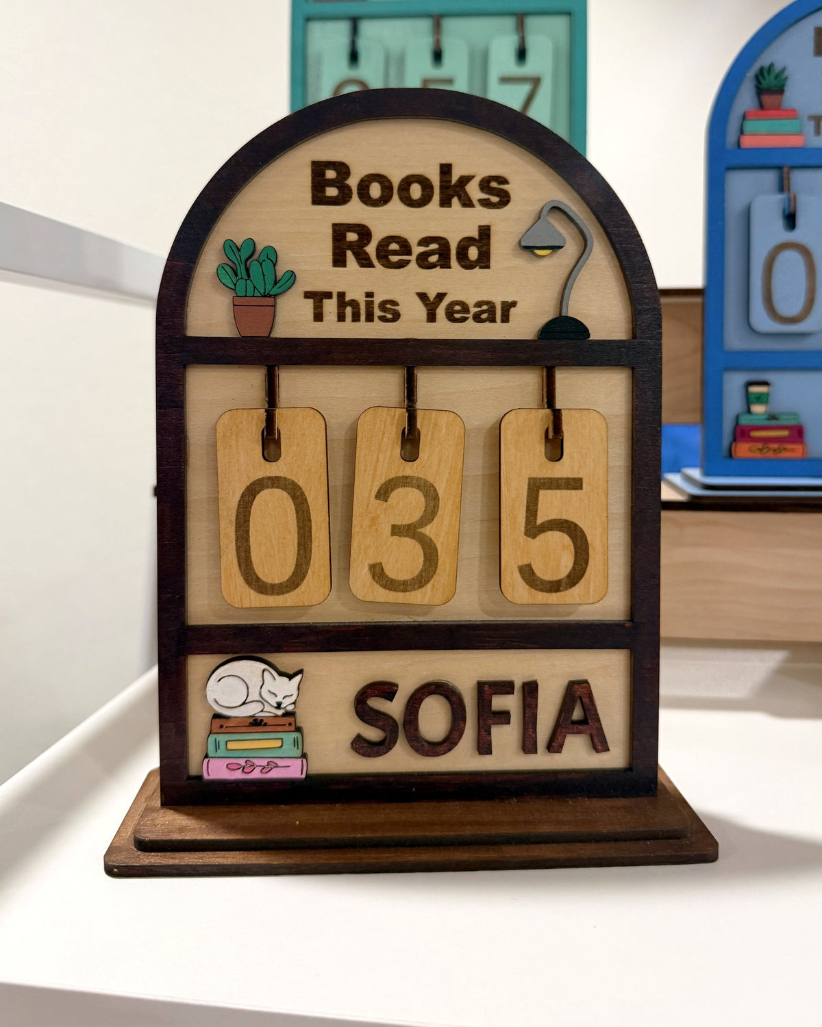 Souvenir Design Books Read This Year wooden counter in brown, number tiles showing 035 and the name SOFIA on the bottom shelf, with more color versions behind it