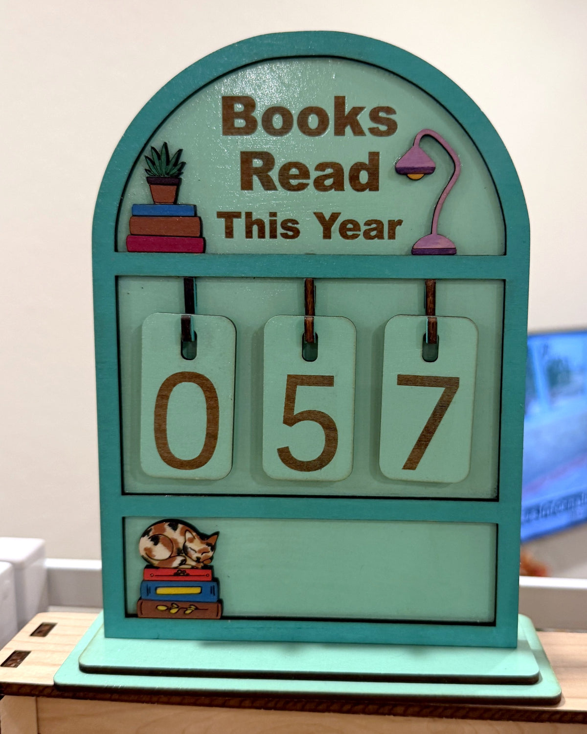 Souvenir Design Books Read This Year wooden counter in mint green, number tiles showing 057 and a calico cat on stacked books