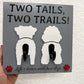 Gray "Two Tails, Two Trails!" leash holder by Souvenir Design with two white dog cutouts seen from behind, two black hooks, red paw prints and "Life's better with two dogs"
