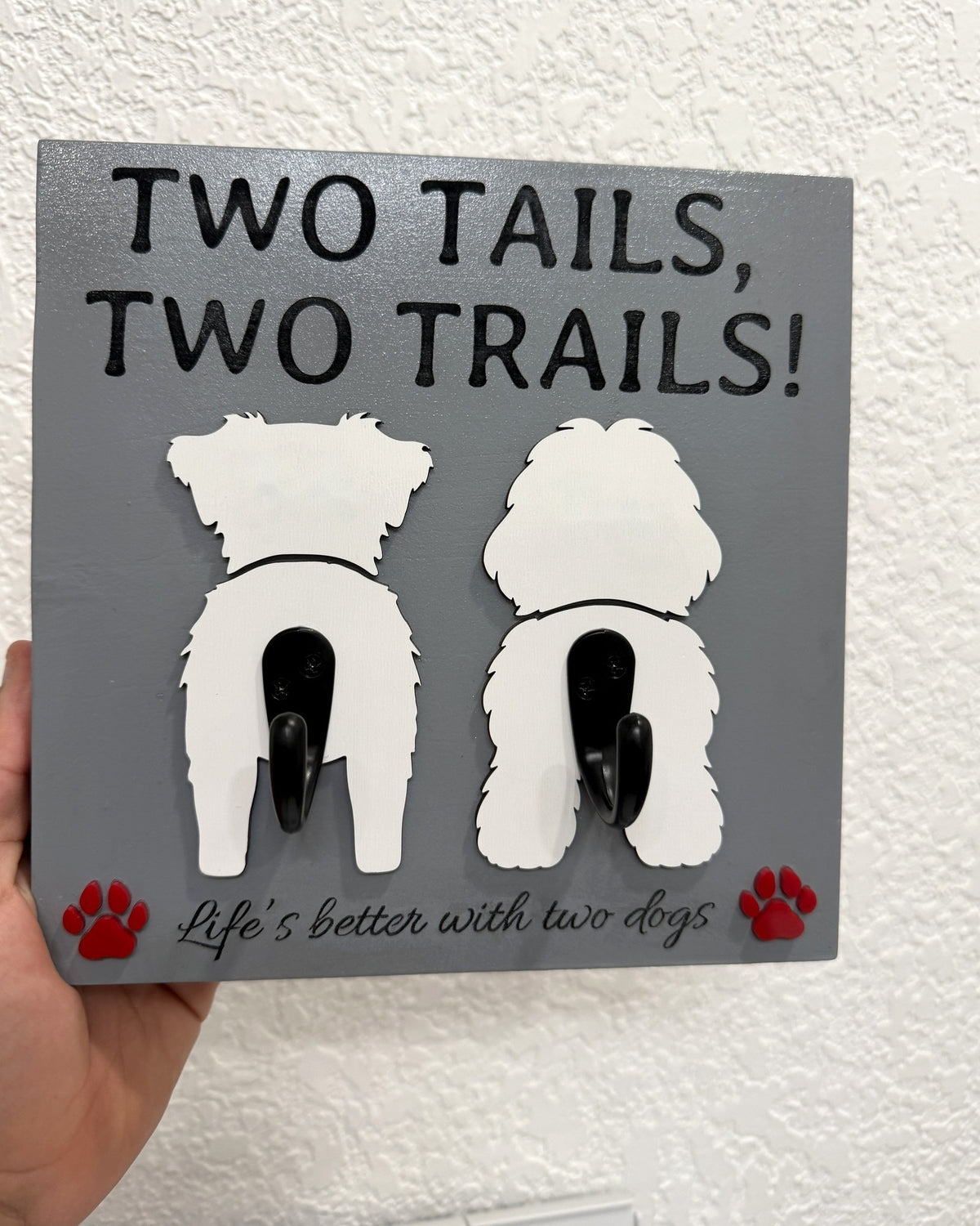 Gray "Two Tails, Two Trails!" leash holder by Souvenir Design with two white dog cutouts seen from behind, two black hooks, red paw prints and "Life's better with two dogs"
