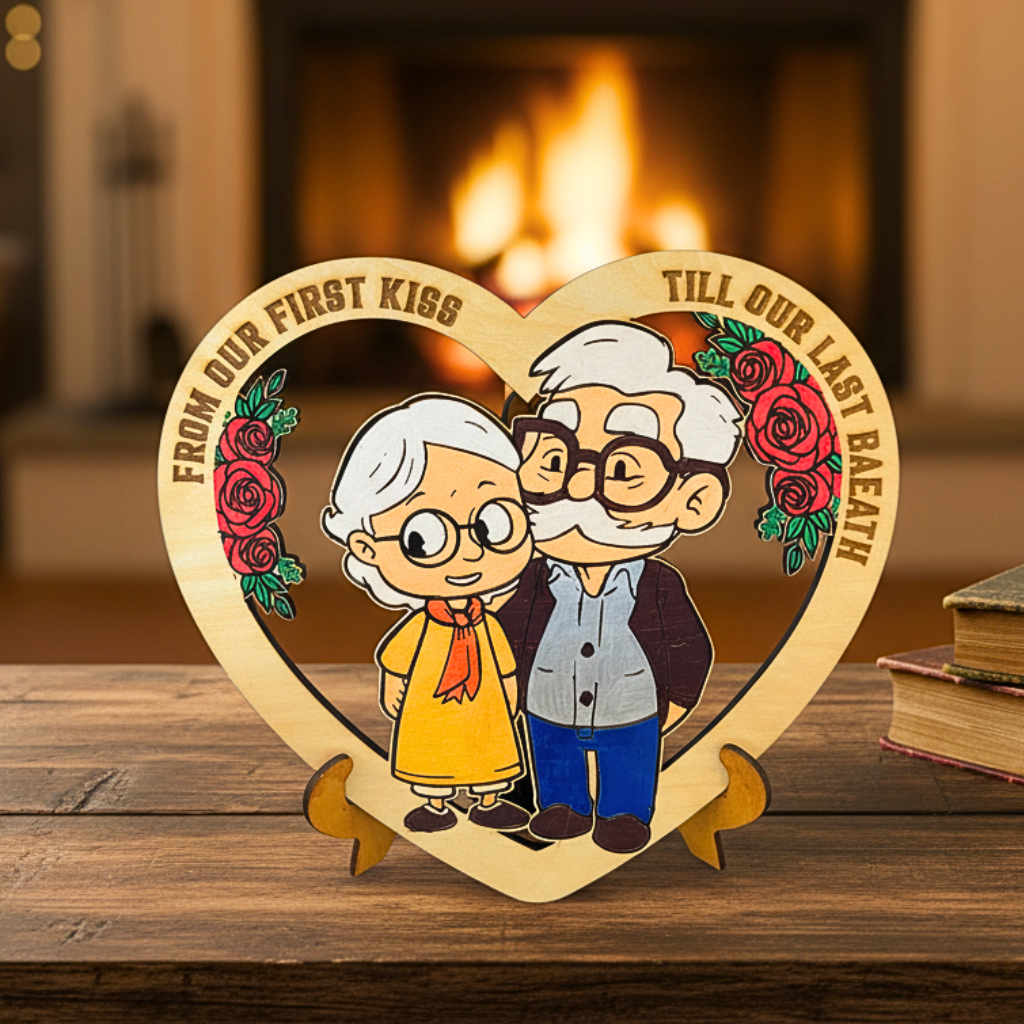 Heart-shaped wooden plaque with cartoon illustration of an elderly couple and romantic text, placed on a wooden surface with a fireplace in the background.