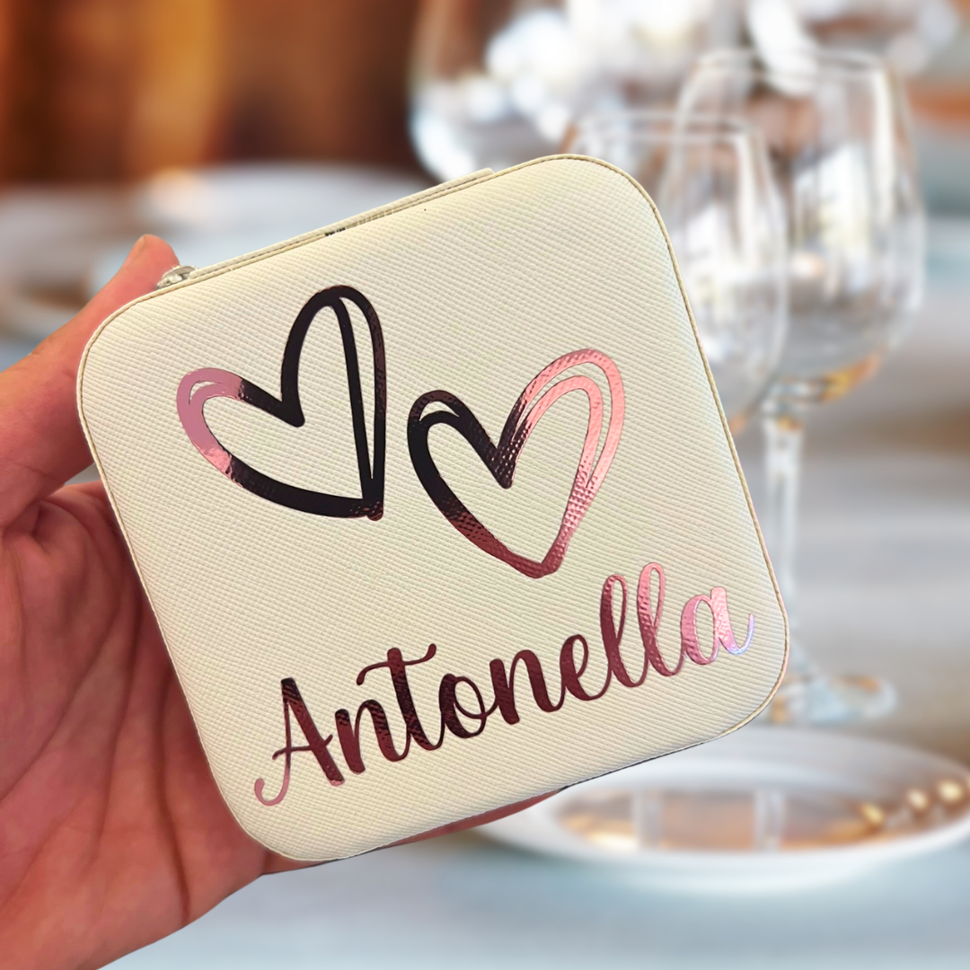 Beige jewelry box with 'Antonella' and heart designs held in a hand with a blurred background of glasses and plates.