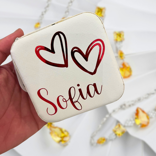 Small white pouch with red hearts and 'Sofia' text held by a hand, with blurred yellow flowers in the background.