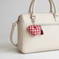 Beige handbag with a red and white checkered heart charm on a light gray background